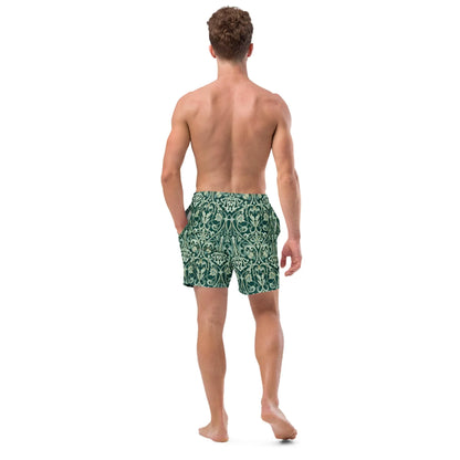 A shirtless man stands barefoot, facing away, with one hand in the pocket of his dark green Oz Swim Trunks by Printful, featuring a light ornate pattern and four-way stretch, water-repellent fabric.