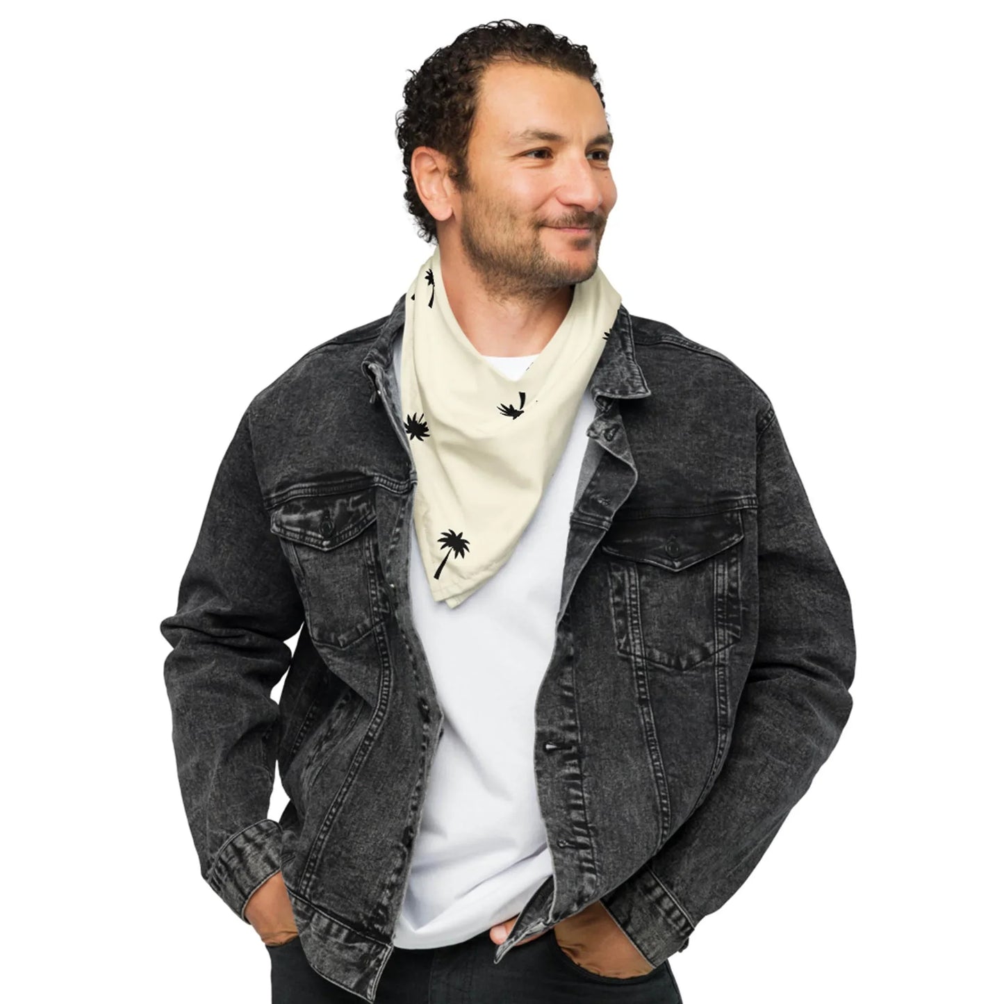 A man wearing a black denim jacket over a white shirt smiles while looking to the side. He has short curly hair and wears a Mambí-inspired light-colored scarf with small black palm tree prints.