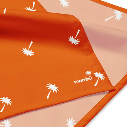 An orange fabric with white palm tree prints and a stitched edge, partially folded to reveal a lighter orange underside with the same pattern and the word Mambí printed in white in the corner.