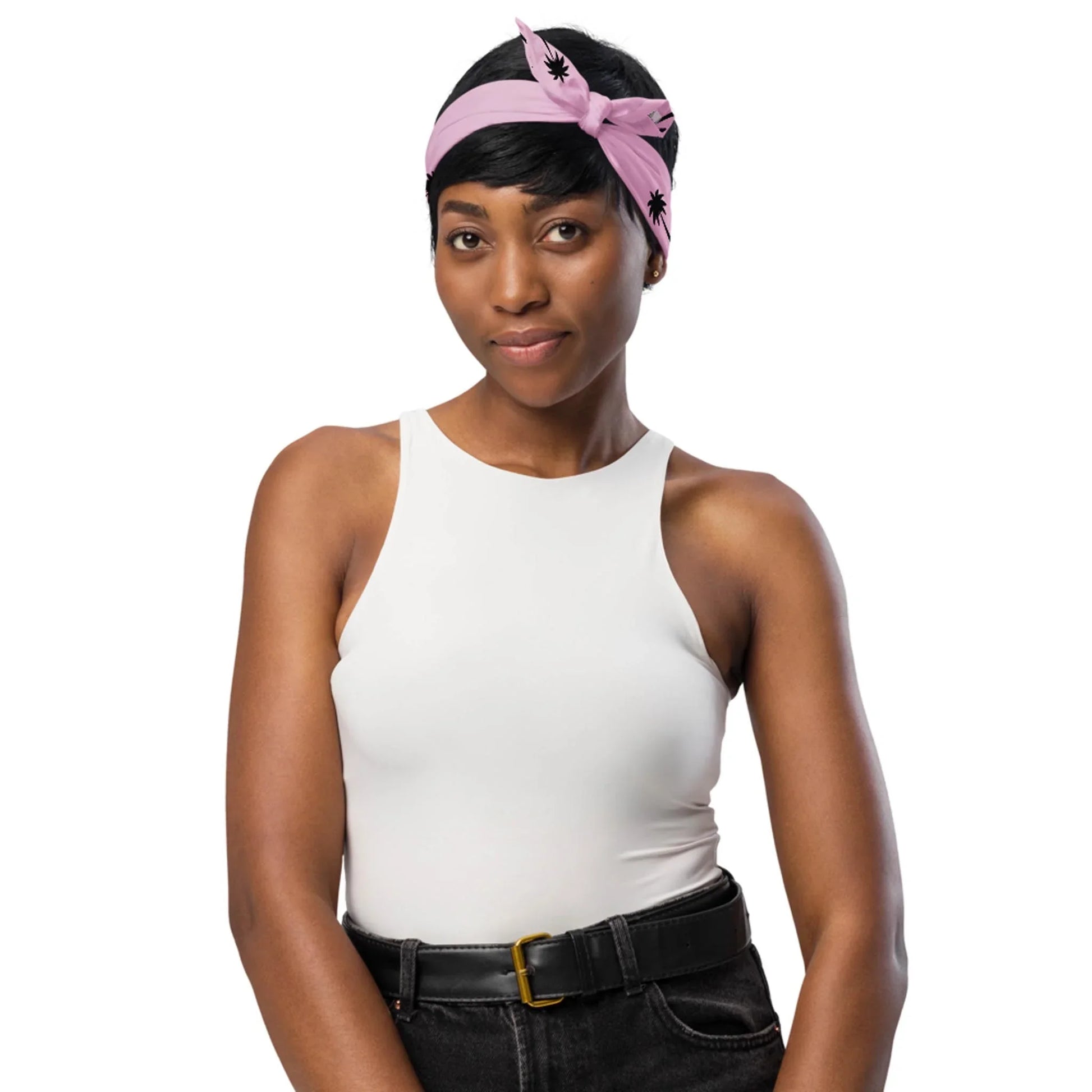 A woman wearing a sleeveless white top, Mambí black high-waisted jeans with a belt, and a pink headband with black designs stands against a white background, smiling softly.