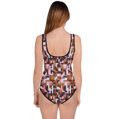 A girl with long brown hair faces away, wearing the Printful “Pixie” One-Piece Swimsuit for big kids and teens. The suit features a colorful pixelated pink, brown, black, and white pattern and UPF 50 sun protection against a plain white background.