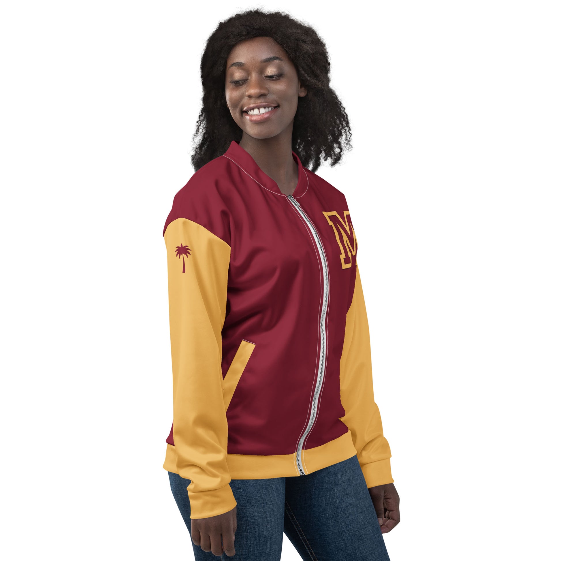 A woman smiles with her eyes closed, wearing a maroon and yellow varsity bomber jacket with an M on the chest and a small palm tree on the sleeve. She has curly hair and stands against a plain white background.
