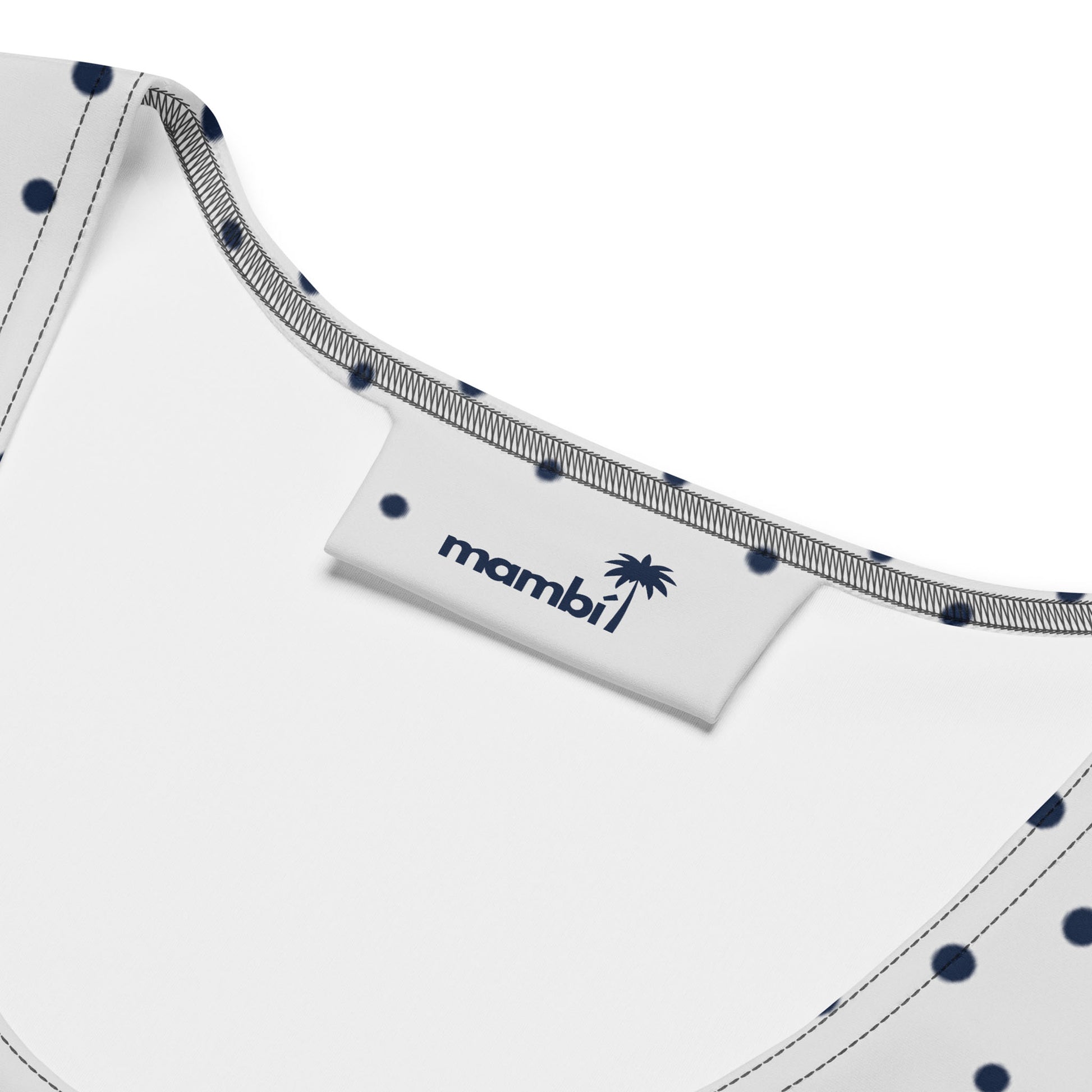 Close-up of the inside neckline of an all-over printed dress with white fabric, navy blue polka dots, and gray stitching. A white tag displays the brand name “mambi” with a palm tree graphic in navy blue.