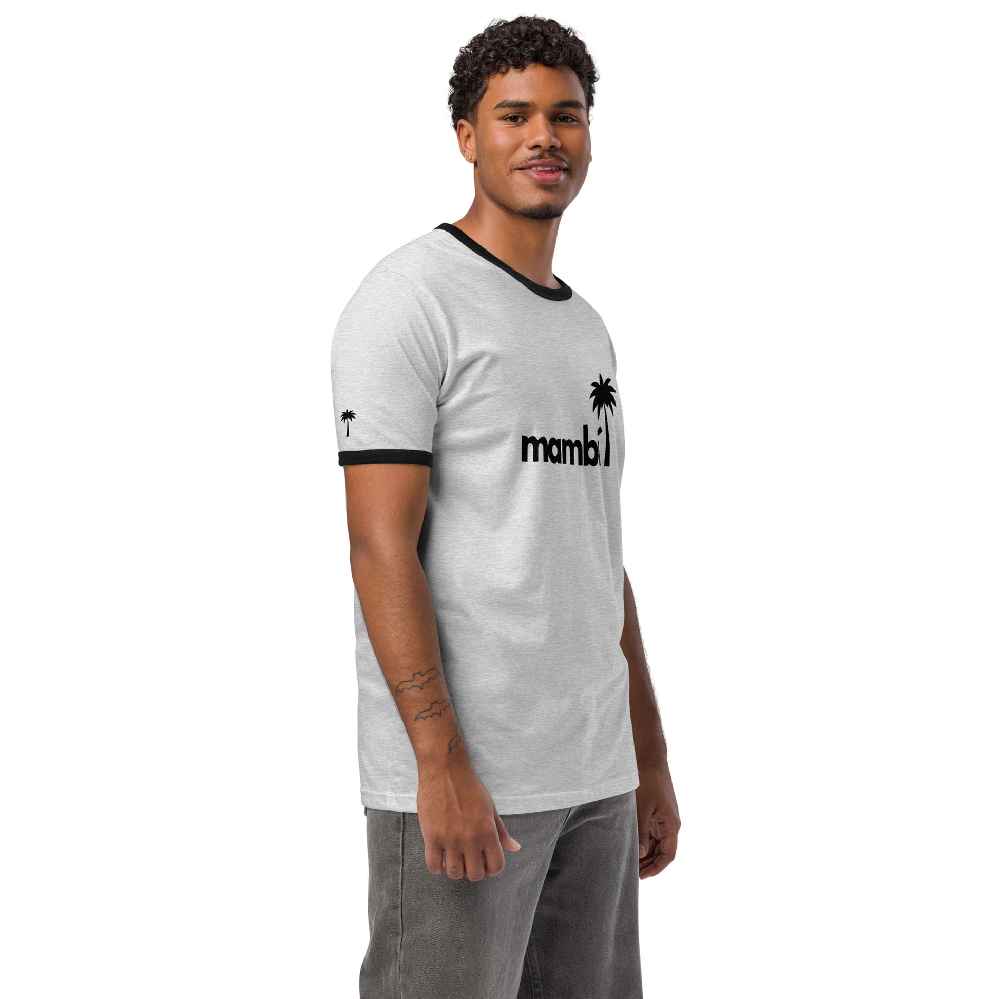 A man with short curly hair and a trimmed beard wears a relaxed fit, lightweight cotton tee with black collar and sleeve hems. The shirt features mambi with a palm tree graphic, giving it a retro look. He stands smiling against a white background.