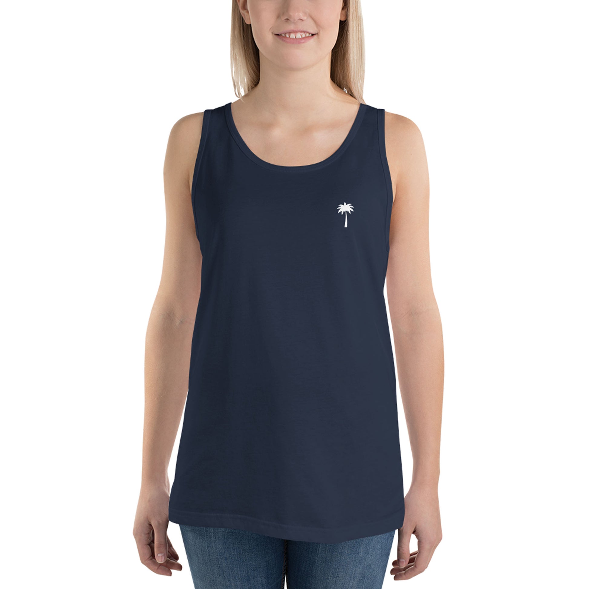 A woman wearing a sleeveless navy blue classic tank top made from ringspun cotton, featuring a small white palm tree graphic on the left chest, paired with blue jeans. She is smiling and facing forward.