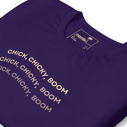 A close-up of a purple graphic tee with the words “CHICK, CHICKY, BOOM” repeated three times in bold, light-colored text. The comfortable cotton shirt’s label at the collar displays the brand name “mambí.”.
