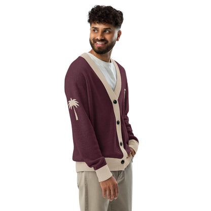 A man with curly hair and a beard smiles while wearing a maroon lightweight knit unisex sweater with beige trim and a palm tree design on the sleeve, a white T-shirt, and beige pants against a white background.
