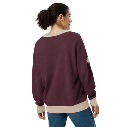 A woman with curly hair in a ponytail wears a loose, maroon two-tone cardigan with beige cuffs and hem, paired with blue jeans. She faces away and smiles, standing against a plain white background.