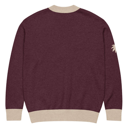 A maroon unisex sweater in a lightweight knit, featuring beige ribbed cuffs, collar, and hem. A partial beige palm tree graphic decorates the left sleeve near the shoulder. Sweater is shown from the back.