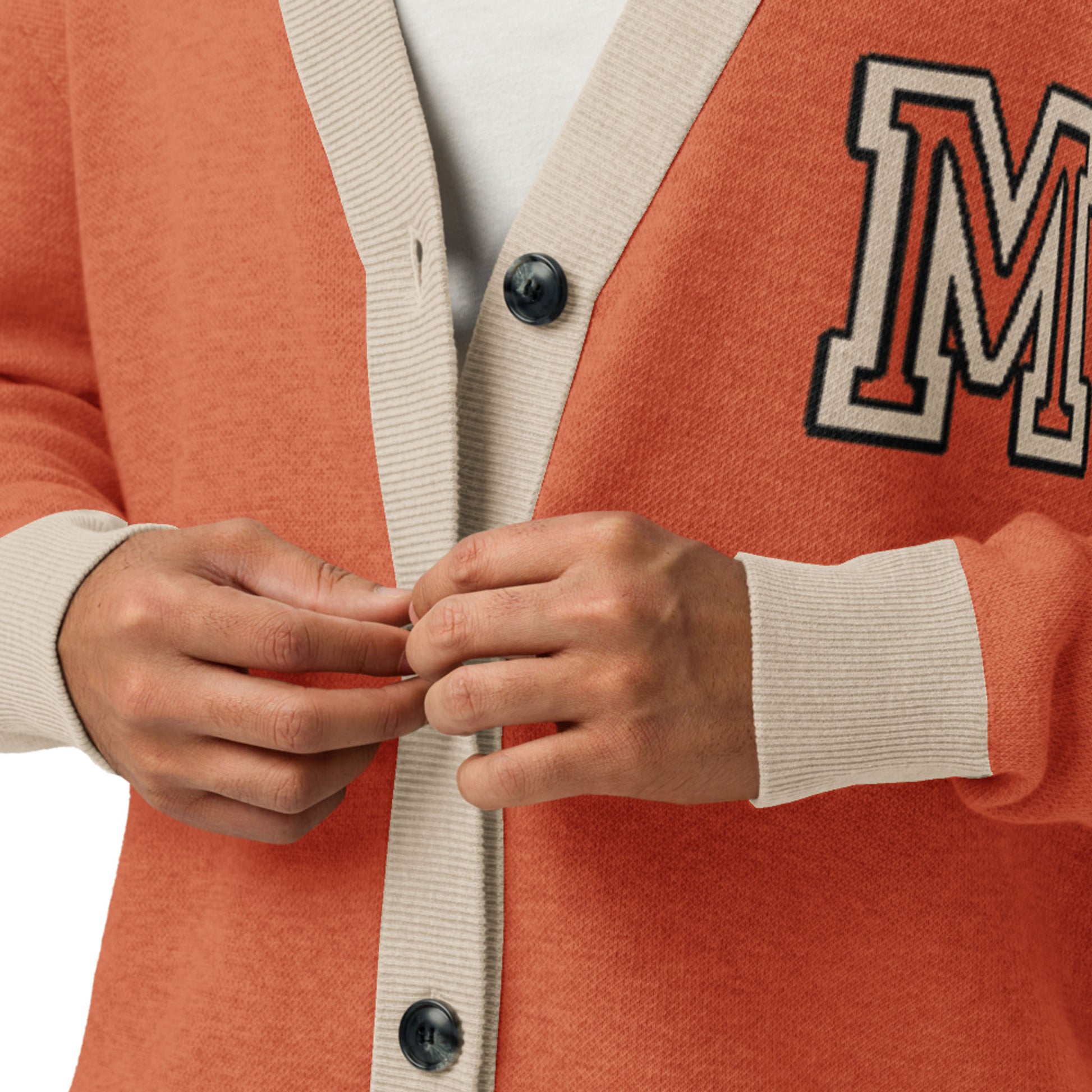 A person wearing a two-tone cardigan—orange with light beige trim and large black buttons—adjusts the front. The unisex cardigan features a bold patch with the letter M on the left side.