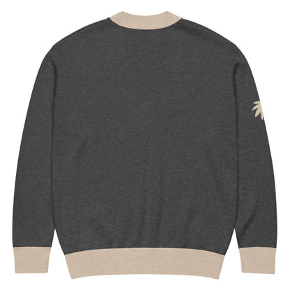 A dark gray two-tone cardigan with light beige ribbed cuffs, hem, and collar. Shown from the back, this unisex cardigan features a small, light beige palm tree design on the upper left sleeve.