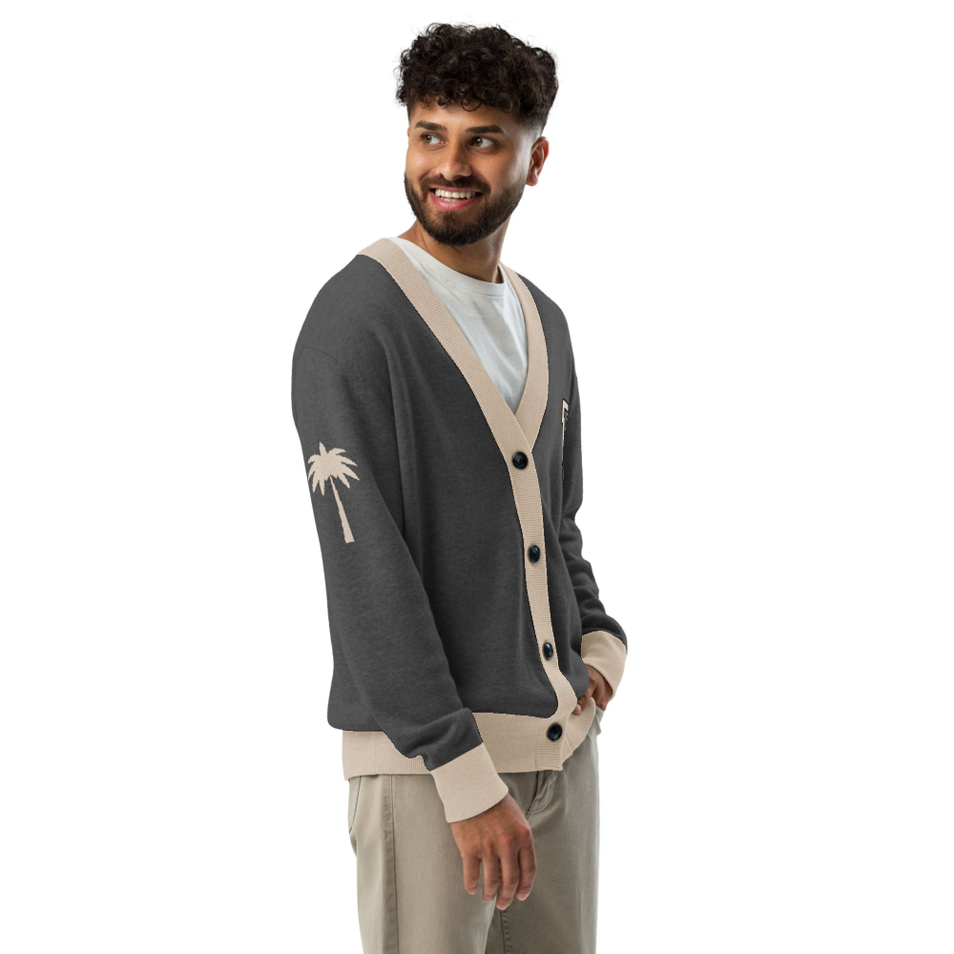 A man with curly hair and a beard smiles while wearing a two-tone unisex cardigan featuring beige trim and a palm tree design on the sleeve, layered over a white shirt and beige pants, standing against a white background.