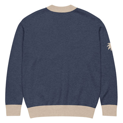 A navy blue jacquard knit sweater with beige ribbed cuffs, collar, and waistband. There is a small beige palm tree graphic on the left sleeve near the shoulder. The soft cardigan is shown from the back.
