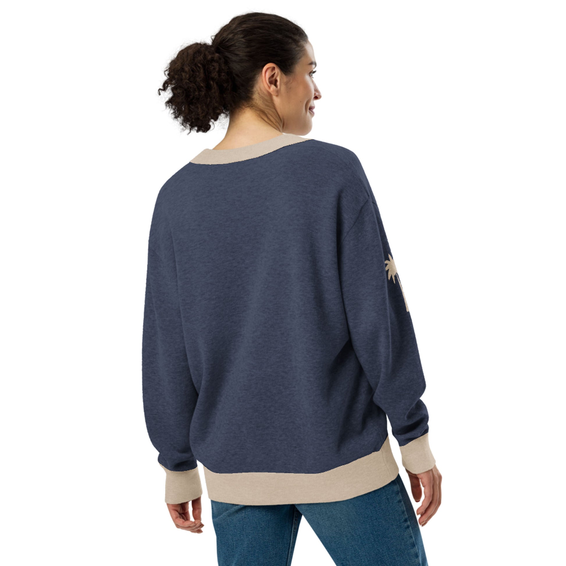 A woman with curly hair in a ponytail is wearing a loose, two-tone cardigan with beige cuffs and hem, paired with blue jeans. She is standing and facing away, looking over her shoulder.