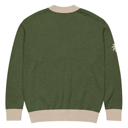 A green unisex cardigan with beige ribbed cuffs, collar, and hem. A small, beige palm tree graphic accents the left sleeve near the shoulder. The image shows the back of this stylish two-tone cardigan.