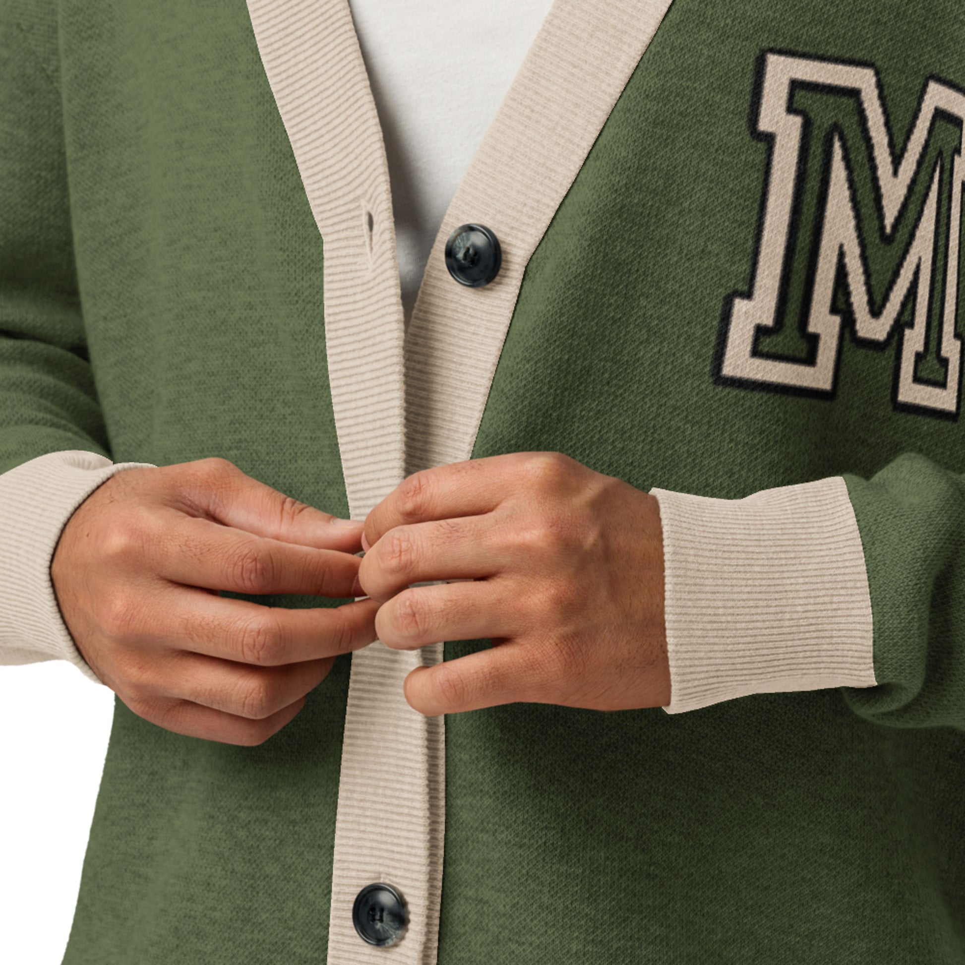 A person fastens a green two-tone cardigan with beige trim and buttons. The left side features a large, black-outlined letter M patch. Only the hands and torso are visible in this stylish unisex cardigan.