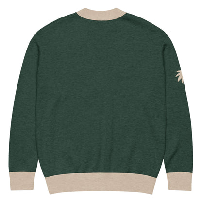 A dark green two-tone cardigan with beige cuffs, hem, and collar, shown from the back. A small beige palm tree graphic appears on the left sleeve.