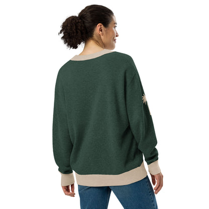 A woman with curly hair in a ponytail is wearing a two-tone cardigan with beige cuffs and hem, along with blue jeans. She is standing and facing away, looking slightly to the side.