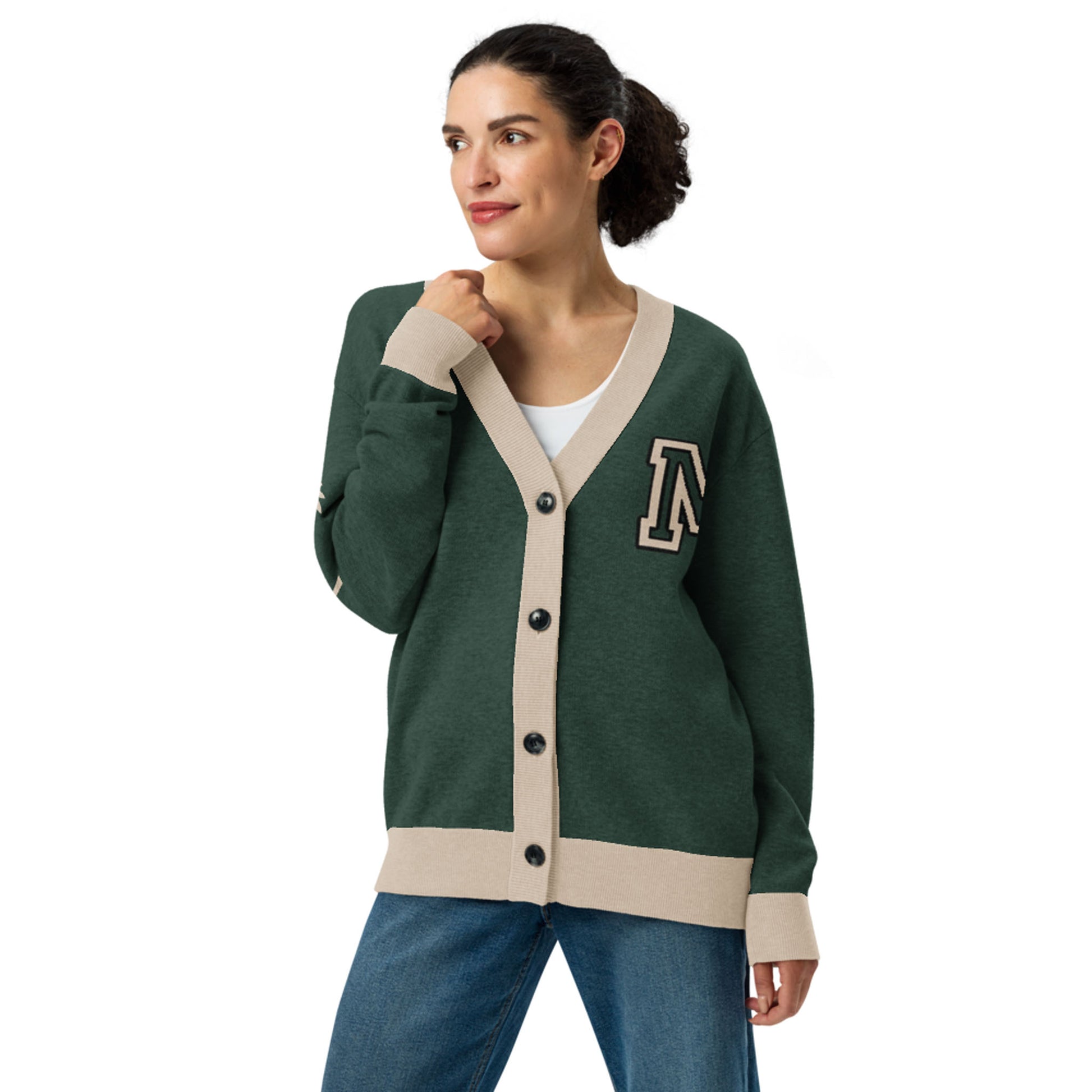 Woman wearing a green two-tone cardigan with beige trim and black buttons, featuring a large letter A patch on the left chest, white shirt underneath, and blue jeans, standing and looking to the side with a slight smile.