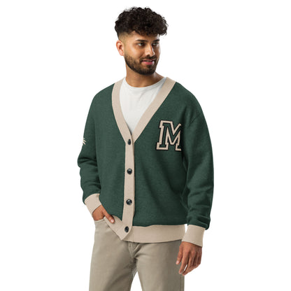 A man with curly hair and a beard wears a green and beige two-tone cardigan with a large M patch, a white T-shirt, and beige pants. He stands with one hand in his pocket, looking to the side and smiling.