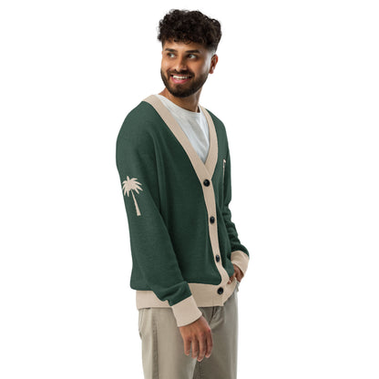 A man with curly hair and a beard wears a unisex two-tone cardigan featuring a palm tree design on the sleeve, paired with a white t-shirt and beige pants. He smiles warmly while looking to the side.