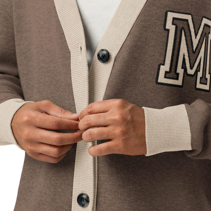A close-up of a person buttoning a two-tone cardigan with large buttons and an embroidered letter M patch on the chest. This unisex cardigan, worn over a white shirt, features stylish brown and beige colors.