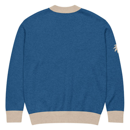 Blue jacquard knit sweater with beige cuffs, neckline, and hem. The back is plain, and there is a small palm tree graphic on the right sleeve near the shoulder. A soft cardigan for a unisex look.