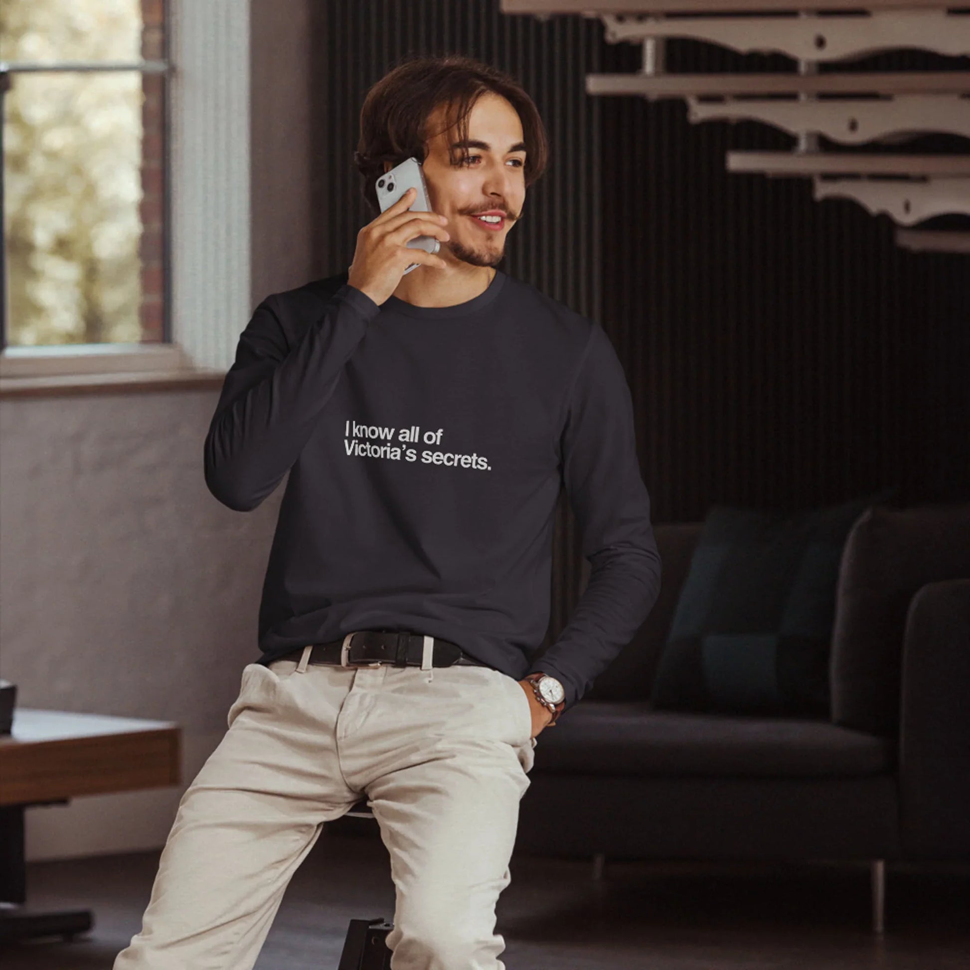 Man wearing a black long-sleeve graphic t-shirt with text, standing in a room with dark walls and a window.