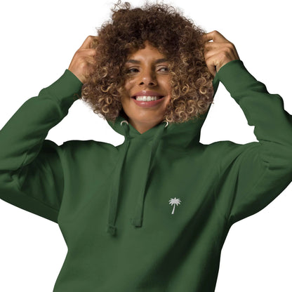 A smiling person with curly hair wears a dark green Mambí hoodie with a small white palm tree logo on the chest and holds the hood with both hands against a white background.