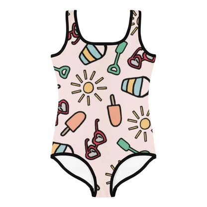 A one-piece swimsuit by Mambí with a light pink background featuring playful illustrations of suns, popsicles, heart-shaped sunglasses, beach balls, and green shovels, all outlined in black.