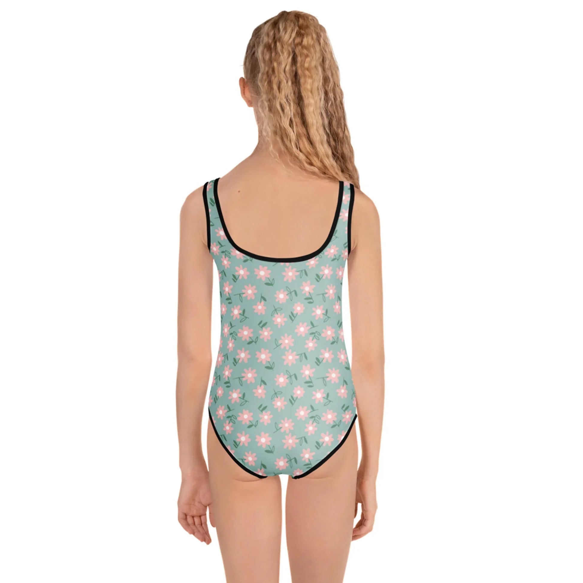 A girl with long, wavy blonde hair faces away from the camera, modeling the Printful Sara One-Piece Swimsuit for toddlers and little kids in light green with bright pink and white daisy prints and black trim against a plain white background.