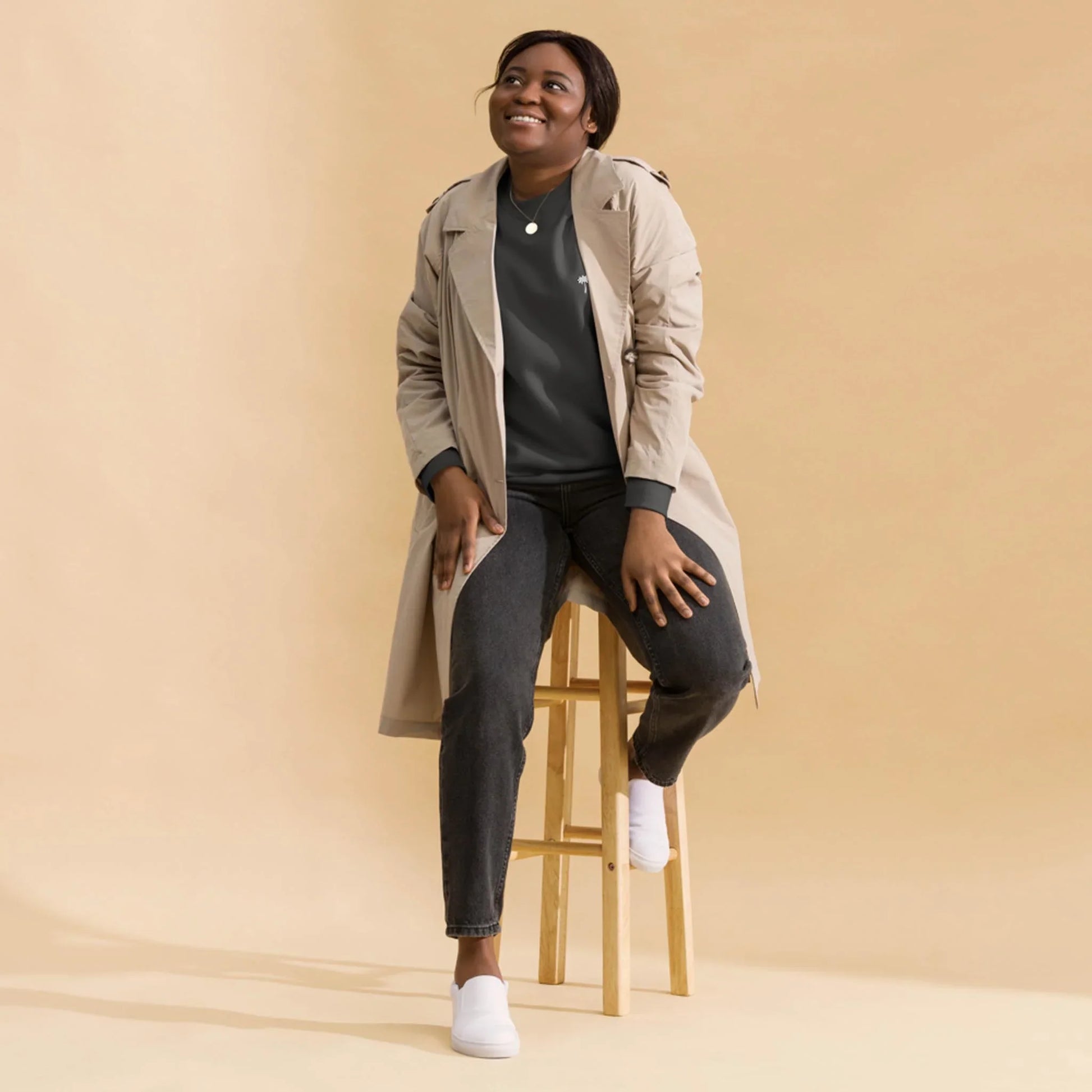 A person wearing a beige Mambí trench coat, dark jeans, and white shoes sits on a wooden stool against a plain beige background, smiling and looking slightly upward.