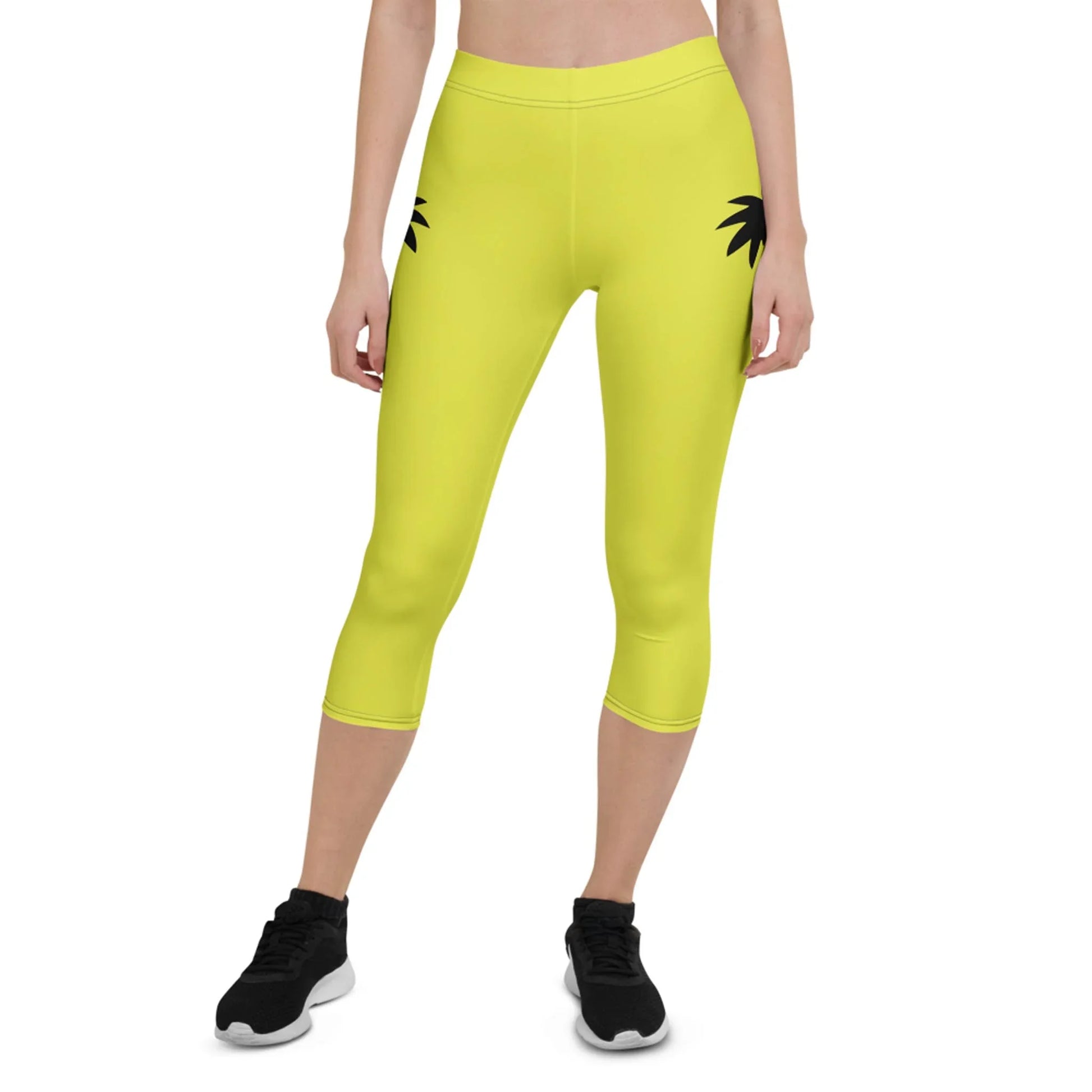 A person wearing bright yellow Mambí capri leggings with black palm leaf prints near the hips, paired with black sneakers. The image shows the lower half of their body against a white background.