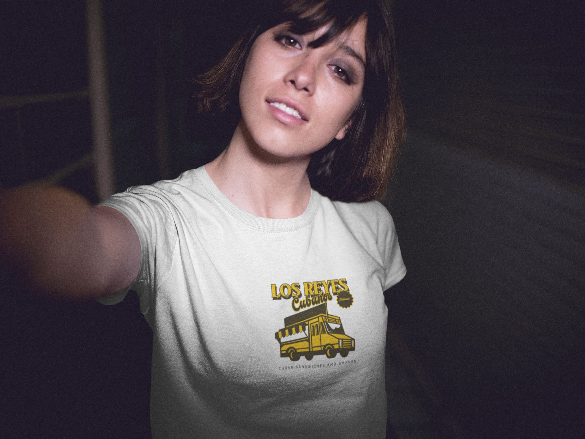 Woman wearing a white graphic t-shirt with 'Los Reyes Cubanos' logo, taking a selfie.