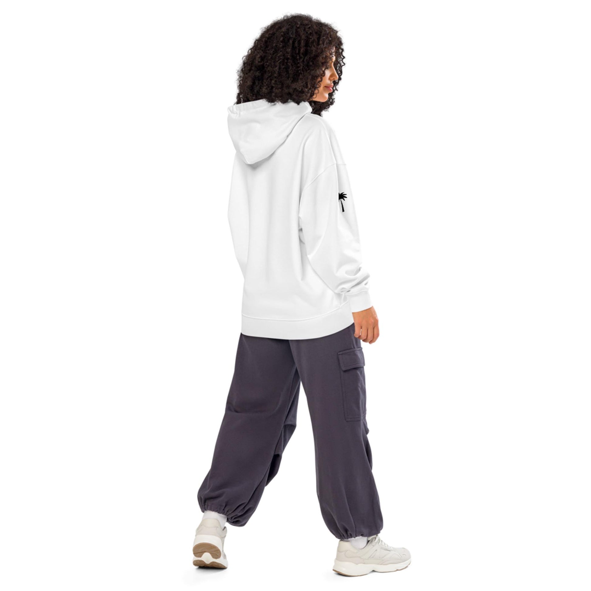 A person with curly hair wears a white Mambí hoodie and dark gray cargo pants with white sneakers, standing and looking to the side against a plain white background.