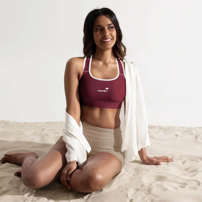 A woman sits on sand wearing a maroon Mambí sports bra with white trim, cream high-waisted shorts, and a loose white shirt draped over her shoulders, smiling and looking slightly to the side in a bright studio setting.