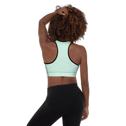 Woman with curly hair wearing a light mint green Mambí sports bra and black leggings, standing with her back to the camera and one hand resting on her head.