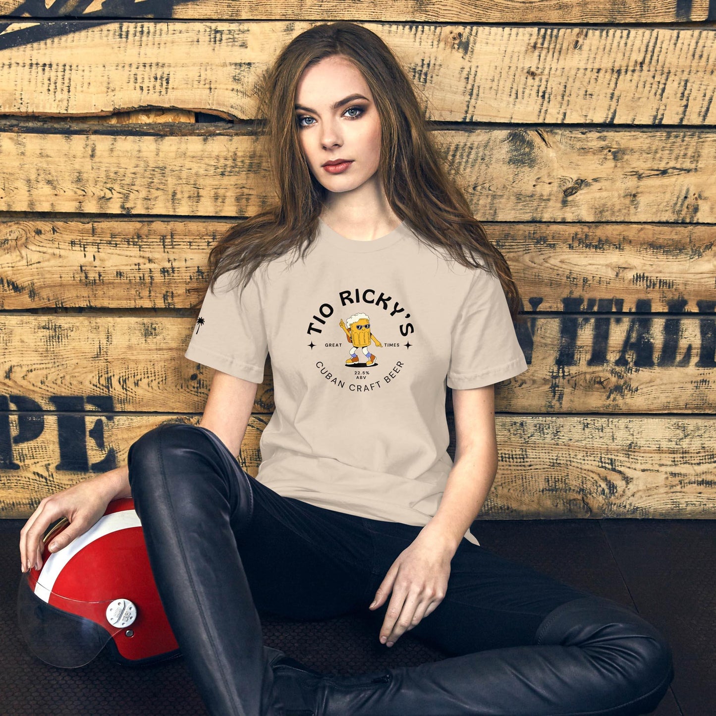 A woman with long brown hair sits on the floor against a wooden wall, wearing a light-colored Mambí Cuban Craft Beer T-shirt and black leather pants, with a red and white motorcycle helmet beside her.