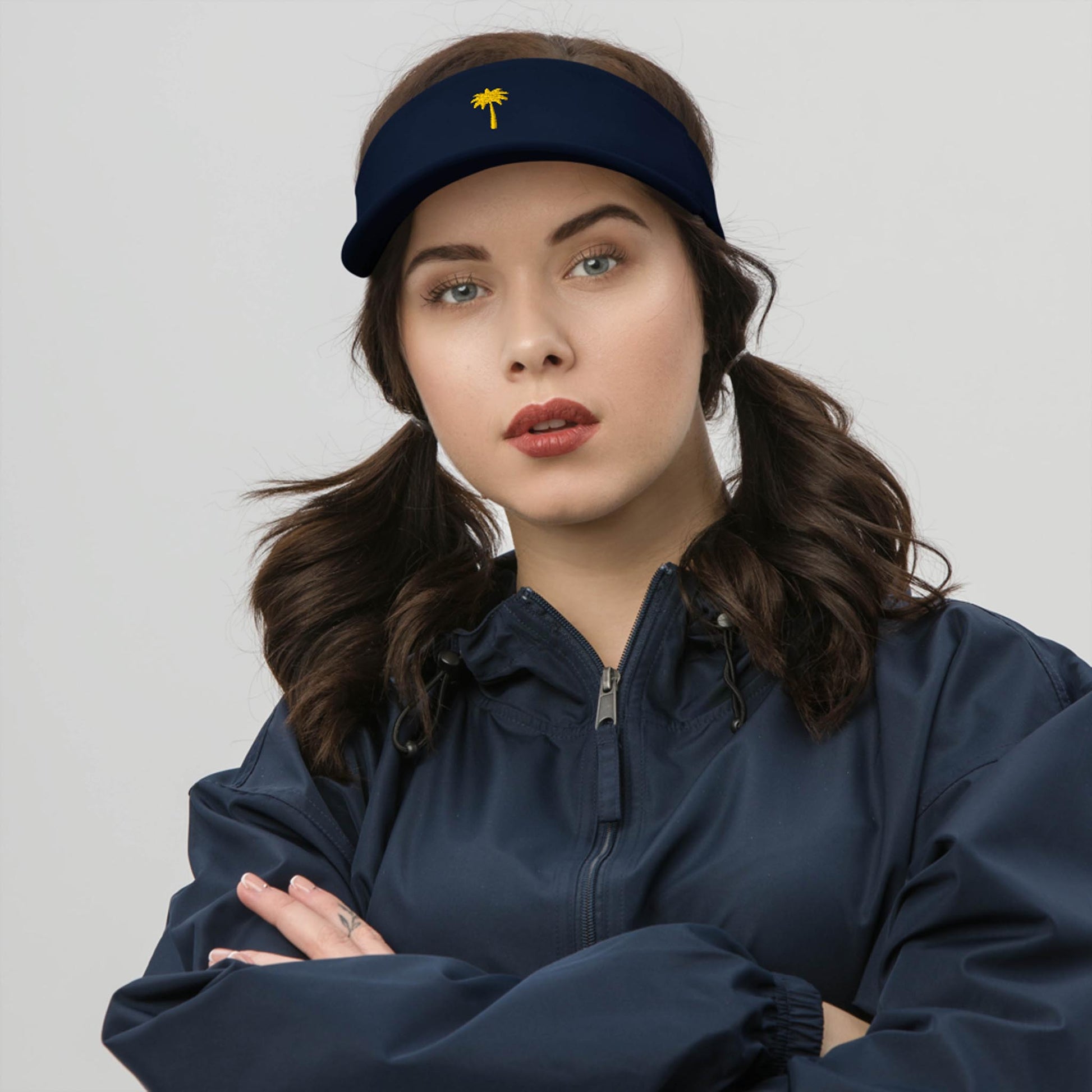 A woman with dark brown hair in pigtails wears a navy visor featuring a small yellow palm tree emblem and a matching Mambí navy jacket, standing with folded arms against a plain light background.