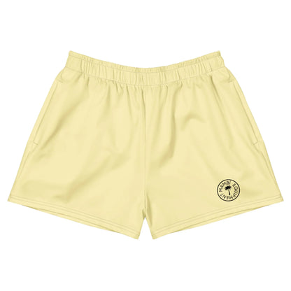 Light yellow athletic shorts with an elastic waistband and a small round black logo near the bottom left hem that reads Mambí Miami Club Wear, featuring a sun icon in the center.
