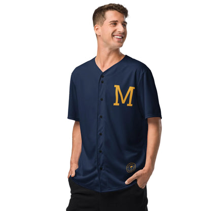A smiling man stands hands in pockets, wearing the Printful Zoilo Classic Baseball Jersey | Unisex in navy blue, featuring a large yellow M on the chest and a round patch near the hem.